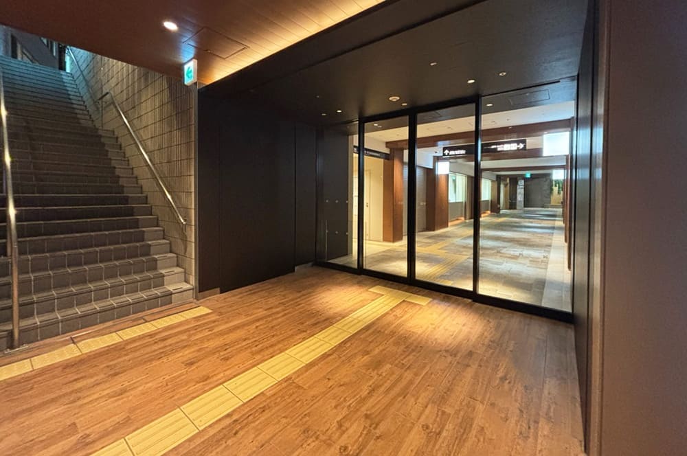 The entrance to the underground passage, featuring wood-grain flooring. Stairs are visible on the left, and beyond the glass door straight ahead, a corridor extends further into the building.