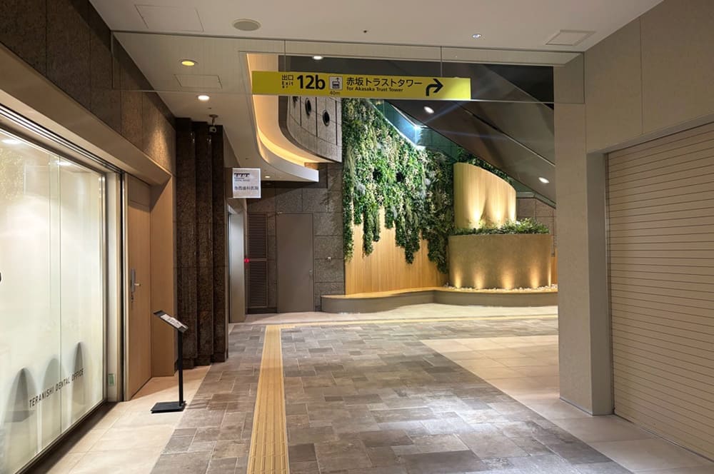 A spacious area at the end of the underground passage. Beyond the overhead sign for "Exit 12b Akasaka Trust Tower," a wall is covered with green foliage, and an escalator is visible in the back right.
