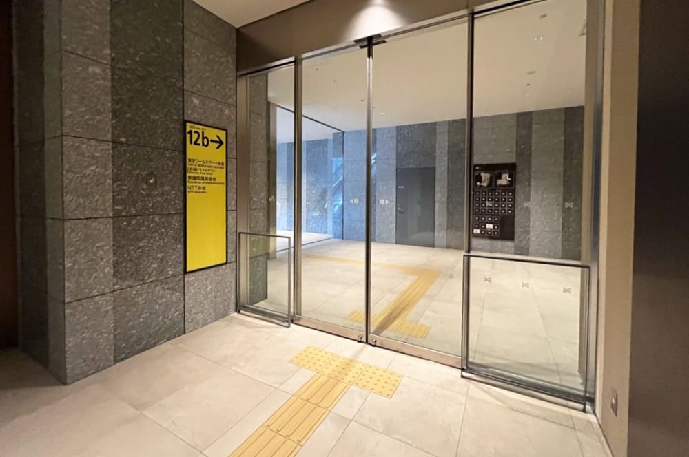 Near Exit 12b of Tameike-sanno Station, featuring stone walls and a glass automatic door. A yellow sign reads "12b Tokyo World Gate Akasaka," and a spacious hall is visible beyond the door straight ahead.