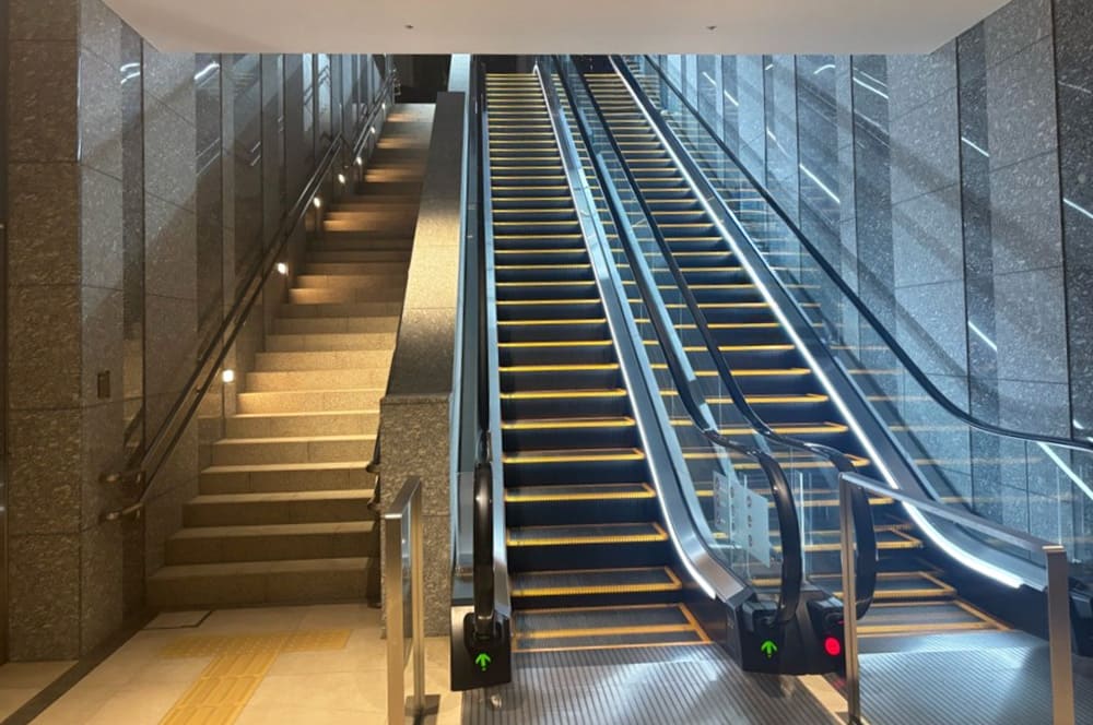 Modern escalators and stairs surrounded by stone walls and lighting. Two upward escalators are on the right, while illuminated stairs run along the left side.