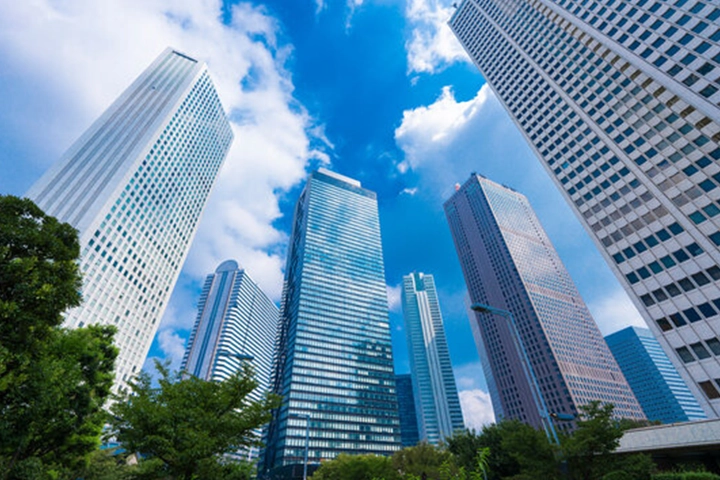 A low-angle view of the high-rise buildings at Shinjuku West Exit, soaring into a blue sky with white clouds.