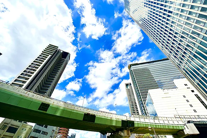 A dynamic low-angle composition looking up at the skyscrapers in the Higobashi area, with an elevated overpass in the foreground against a backdrop of vibrant blue sky and white clouds.