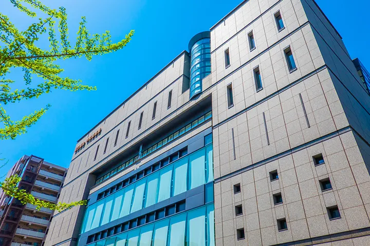 Modern exterior of the Osaka City Central Library building against a bright blue sky with fresh green leaves in the foreground.