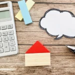 A calculator, a sheet of paper with speech bubbles, and a small wooden house
