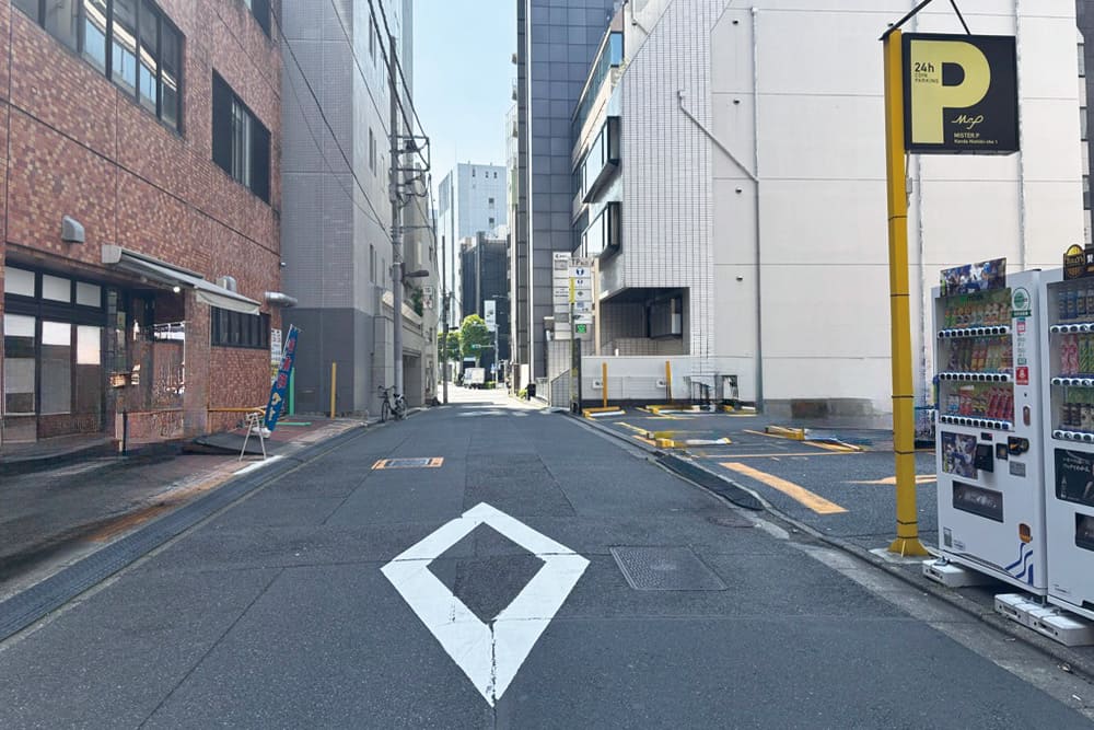 一方通行の道路。白の実線と菱形の標示、右手に24時間営業の駐車場を示す看板。