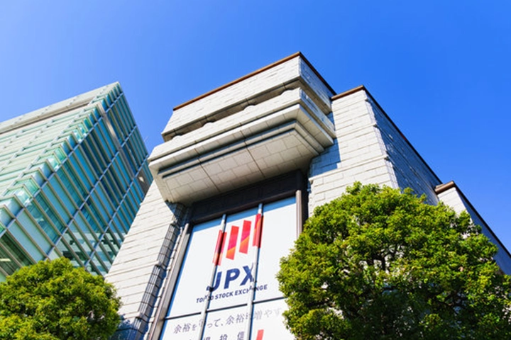 青空の下、東京証券取引所の建物を見上げた構図。モダンなガラス張りのビルが隣接している。
