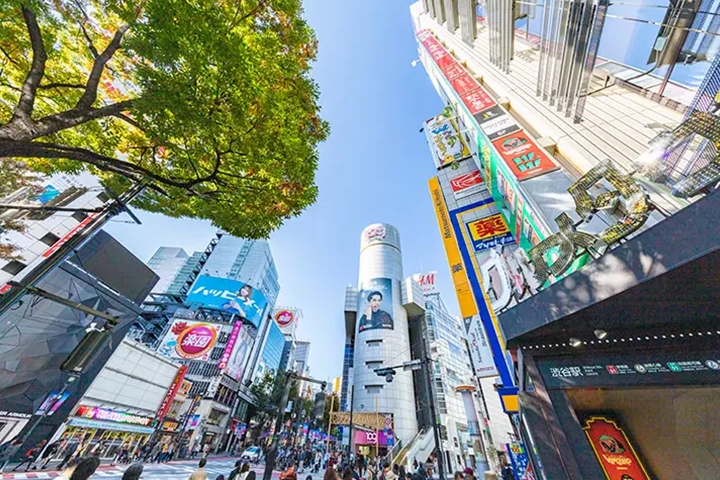 渋谷の駅前。高層ビル群に大型ビジョンや看板が並び、多くの人々が歩いている。