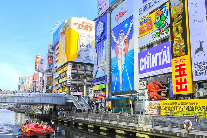 グリコの看板や多彩な広告が並ぶ道頓堀の風景。運河には赤い観光船が浮かび、賑やかな街並みを映している。