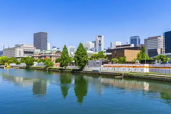 穏やかな川面に街並みが映る中之島の風景。中央には赤レンガ造りの大阪市中央公会堂が見え、その周囲に大阪市役所や近代的な高層ビル群が青空の下に広がっている。