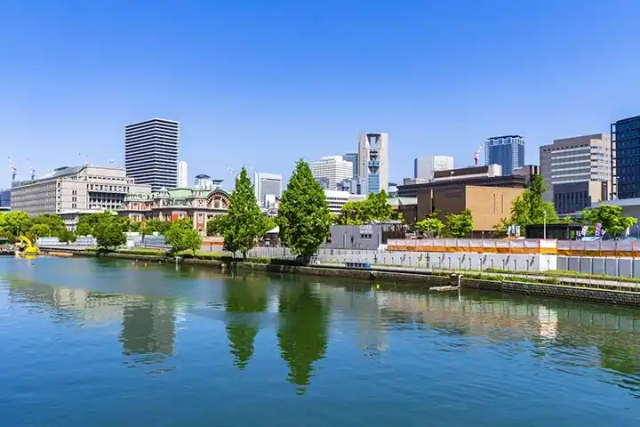 穏やかな川面に街並みが映る中之島の風景。中央には赤レンガ造りの大阪市中央公会堂が見え、その周囲に大阪市役所や近代的な高層ビル群が青空の下に広がっている。