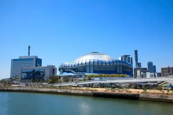 青空を背景にした京セラドーム大阪の全景。特徴的な銀色のドーム形状が目を引き、手前を流れる川の対岸には歩行者用の通路や周辺のビルが並んでいる。
