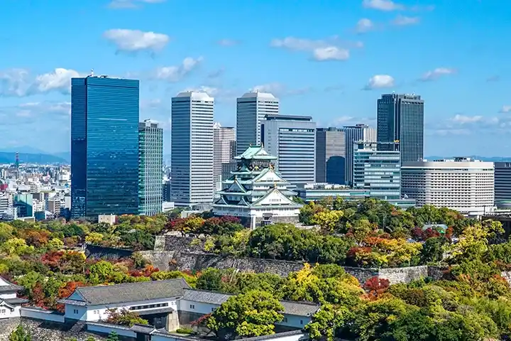 紅葉が始まりかけた木々に囲まれた大阪城天守閣。背景にはクリスタルタワーなど大阪ビジネスパークの近代的な高層ビル群がそびえ立ち、歴史と現代が融合した景色が広がっている。