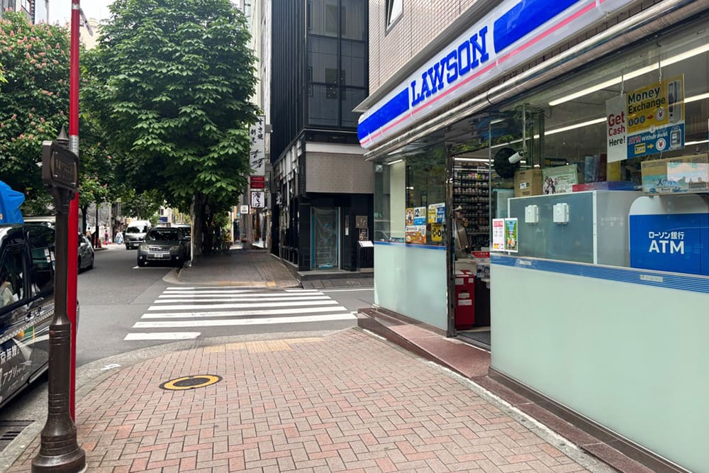 右側にローソンの青い看板と店舗入り口があり、その左側に横断歩道や街路樹が並ぶ銀座の通りが見える風景。