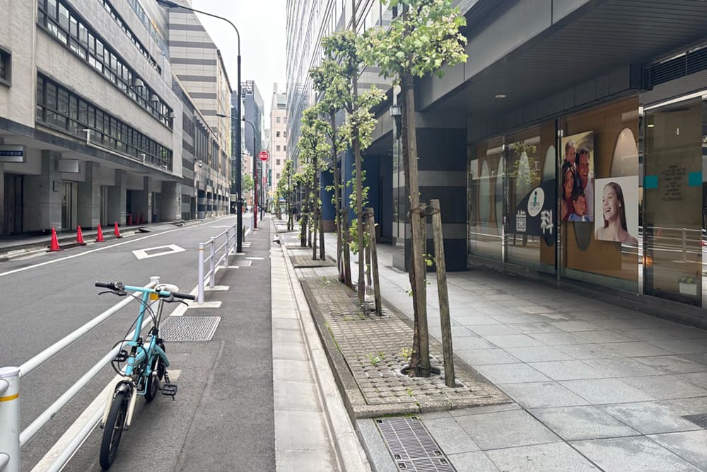 アイタワー歯科クリニックのガラス張りの外観と、街路樹のある歩道および道路の風景。