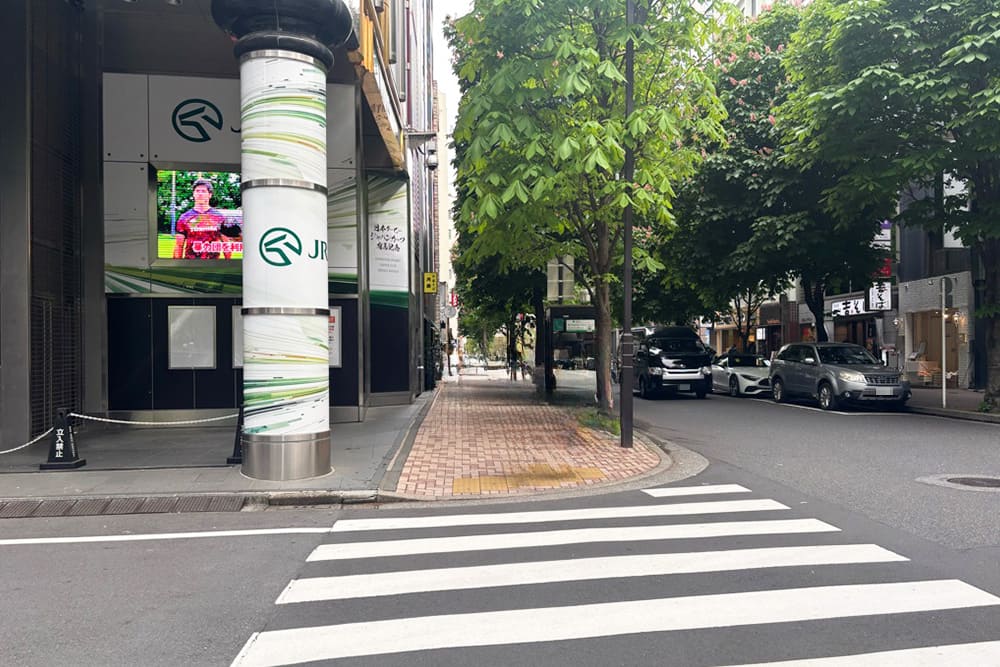 ウインズ銀座のJRAモニターや大型の柱がある角と、手前の横断歩道および奥へと続く街路樹のある歩道の風景。